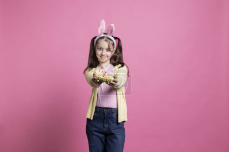 Photo for Sweet young girl confidently smiling for the camera while carrying a basket full of Easter eggs and a chick. Joyful toddler with rabbit ears, pigtails and spring ornaments stands over pink backdrop. - Royalty Free Image