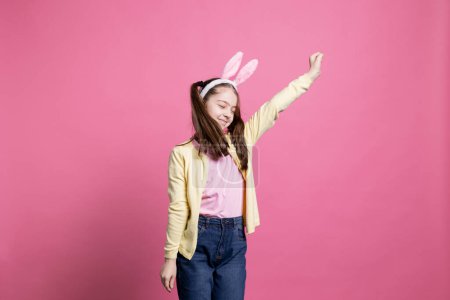 Photo for Positive enthusiastic girl fooling around and dancing on camera, wearing bunny ears in studio and showing dance moves. Young toddler acting cheerful and excited about easter celebration. - Royalty Free Image