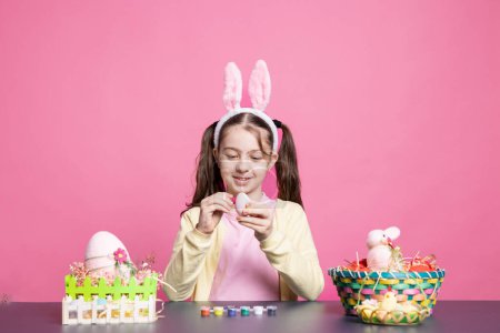 Photo for Cheerful schoolgirl with bunny ears painting eggs at a table, creating handmade festive decorations for easter celebration tradition. Young smiling girl enjoys decorating ornaments with tie dye. - Royalty Free Image