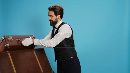 Smiling bellboy carries trolley bags in studio, helping guests with luggage. Professional doorman illustrating role as hotel concierge staff in hospitality industry, tourism concept.