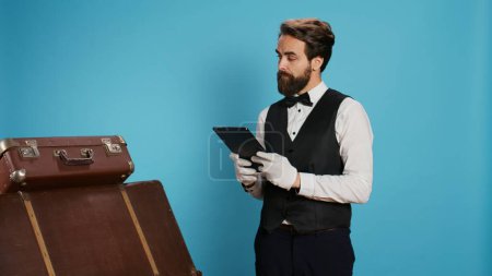 Photo for Classy doorman porter reviewing tablet, in charge of baggage for safekeeping. Bellboy presenting his commitment in providing outstanding concierge service, symbolizing his occupation at chic hotel. - Royalty Free Image
