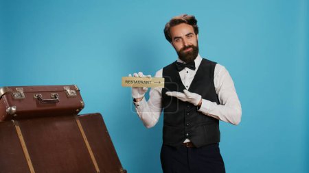Hotel porter advertises restaurant sign to help travellers with premises directions, holding diner pointer while he wears white gloves. Bellhop pointing at symbol for the dining space.