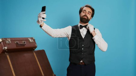 En el estudio, el portero en traje toma fotos mientras usa la aplicación para teléfonos inteligentes para selfies y actúa como un tonto contra el fondo azul. Elegante conserje profesional del hotel haciendo fotos con su teléfono.