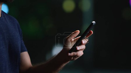 Photo for Modern person on smartphone during stroll late at night, looking around city centre with illuminated skyscrapers. Casual man texting on phone and walking downtown. Handheld shot. Close up. - Royalty Free Image
