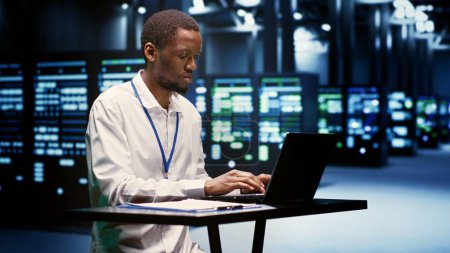 Foto de Hombre de servicio afroamericano evaluando mainframes ayudando a las empresas a administrar bases de datos y almacenar archivos. Electrónica de granja de servidores de inspección profesional utilizada para computación de alto rendimiento - Imagen libre de derechos
