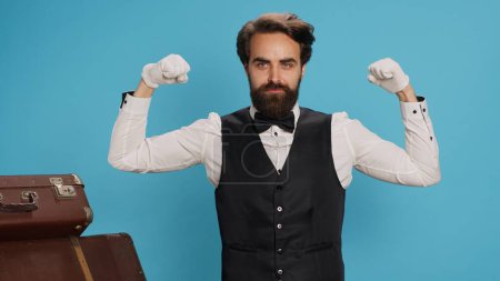 Strong employee flexing arm muscles in studio, indicating power and strength underneath his classy suit and tie. Hotel porter doorman feeling powerful on camera, tourism sector concept.