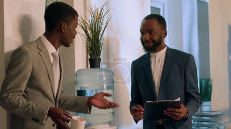 Cheerful african american businessmen enjoying nice water tank conversation during nightshift break. Joyful worker having cup of coffee relaxing while comunicating with colleague in office