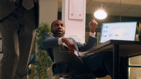 Photo for African american hyperactive employee sneaking up on tired coworker asleep in desk chair during nightshift. Happy businessman shaking up sleepy colleague taking nap in office - Royalty Free Image