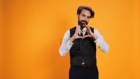 Photo for Butler showing heart shaped symbol in studio, working at five star restaurant and uncovering sincere feelings. Stylish waiter being romantic and presenting his love with sign, honest emotions. - Royalty Free Image