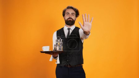 Skilled butler indicates stop sign with palm, posing against yellow background with food tray. Restaurant worker in suit showcasing rejection and dismissal, disagreeing with something.