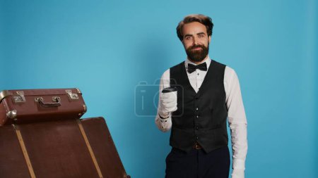 Hotel porter enjoying coffee cup on camera, bellboy wearing professional classy uniform and gloves. Stylish doorman drinking refreshment, posing with luggage on blue background.