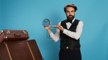 Photo for Classy bellhop poses with wall clock, looking at time and panicking for running late in tourism industry. Punctual hotel concierge employee using watch to see exact hour and minutes. - Royalty Free Image