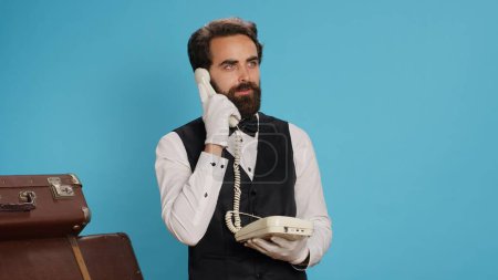 Luxurious bellhop answers landline phone call in studio, talking to travellers about room accommodation. Elegant professional hotel porter using telephone with cord to take calls, vintage gadget.
