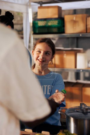 Téléchargez les photos : Portrait d'une jeune femme souriante qui donne gratuitement de la nourriture à une personne dans le besoin dans une banque alimentaire extérieure. Gros plan d'une jeune fille qui apporte joyeusement un soulagement de la faim et un soutien aux pauvres et aux sans-abri. - en image libre de droit