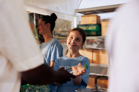 Photo for Cheerful caucasian girl volunteering by handing out free food to the needy at a non-profit food drive event. Kind youngster providing humanitarian aid to homeless people and those in poverty. - Royalty Free Image