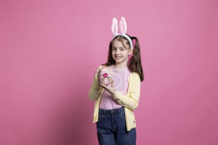 Photo for Small lovely child with bunny ears holding a painted pink egg in front of camera, presenting her handmade easter decoration over colorful background. Young adorable girl shows traditional ornaments. - Royalty Free Image