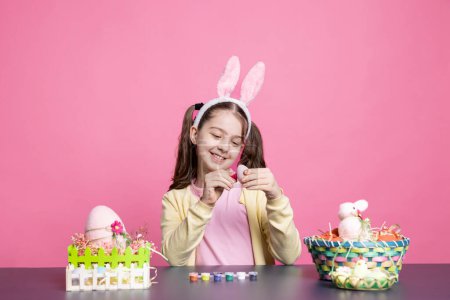 Téléchargez les photos : Joyeux bambin heureux peinture oeufs de Pâques avec aquarelle en studio, décoration ornements festifs faits à la main sur fond rose. Le jeune enfant aime la créativité et l'artisanat, la coloration au pinceau. - en image libre de droit