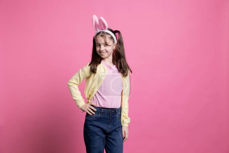 Téléchargez les photos : Doux enfant portant des oreilles de lapin posant devant la caméra, debout sur fond rose. Jeune fille se sentant heureuse et joyeuse, adorable enfant célébrant Pâques temps en studio. - en image libre de droit