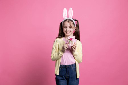Téléchargez les photos : Jeune enfant mignonne posant avec un lapin en peluche rose jouet en studio, présentant ses décorations à temps pour la célébration de Pâques. Petite fille avec des nattes tenant un lapin coloré sur la caméra. - en image libre de droit