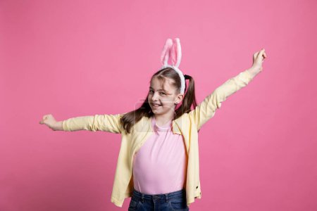 Photo for Joyful small child with pigtails dancing around in front of camera, pointing at something in studio and having fun. Young toddler feeling confident while she is wearing bunny ears. - Royalty Free Image