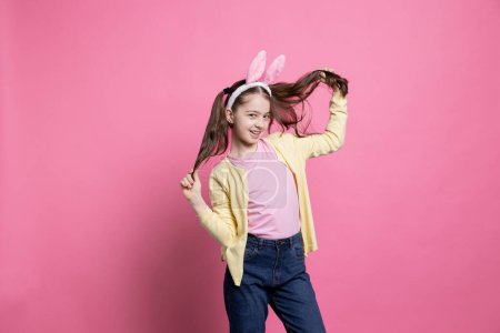 Photo for Smiling confident toddler dancing around in the studio with bunny ears, feeling happy and excited about easter event holiday. Young kid being joyful against pink background, wearing pigtails. - Royalty Free Image