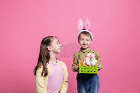 Photo for Cute brother and sister showing festive painted eggs and rabbit toy in a basket, enjoying easter celebration holiday. Little kids siblings posing against pink background, handmade decorations. - Royalty Free Image