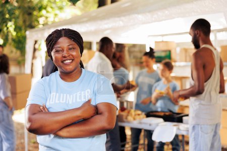 Begeisterte schwarze Frau in blauem T-Shirt mit verschränkten Armen blickt in die Kamera. Portraitaufnahme einer afrikanisch-amerikanischen Freiwilligen, die bereit ist, armen, bedürftigen und weniger glücklichen Menschen humanitäre Hilfe zu leisten