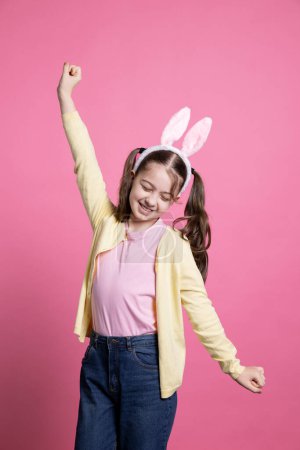 Téléchargez les photos : Joyeux bambin avec des nattes et des oreilles moelleuses qui s'amusent dans le studio, jouant et agissant ridicule devant la caméra. Joyeuse petite fille dansant avec confiance sur fond rose. - en image libre de droit