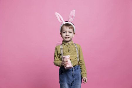 Téléchargez les photos : Doux jeune garçon joyeux posant avec un jouet de lapin moelleux à la caméra, portant des oreilles de lapin et célébrant la fête de Pâques. Petit enfant excité et joyeux sur fond rose. - en image libre de droit