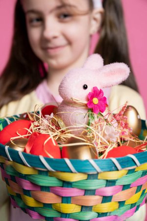Photo for Young cheerful kid holding a colorful decorations basket on camera, showing painted eggs and a stuffed pink rabbit as festive arrangement. Cute girl smiling in studio with adorable decor. Close up. - Royalty Free Image