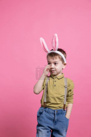 Photo for Joyful young kid feeling confident posing against pink background, wearing bunny ears to celebrate easter holiday event. Small little child smiling in front of camera, waiting to receive presents. - Royalty Free Image