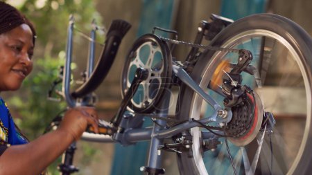Téléchargez les photos : Jeune femme noire sportive tenant et ajustant les composants de vélo dans la cour de la maison. Femme cycliste enthousiaste examinant et réparant soigneusement le vélo endommagé pour le cyclisme de loisirs d'été. - en image libre de droit