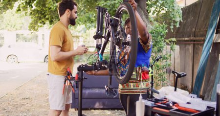 Photo for Committed interracial couple outside servicing bicycle components. Healthy active black woman and caucasian man inspecting and maintaining bike tires and chains for leisure cycling. Pull-back shot. - Royalty Free Image