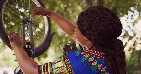 Photo for Healthy black woman mending her own bicycle using specialized equipment from toolkit in yard. Dolly zoom-in shot. Female african american cyclist repairing front bike wheel outdoors. - Royalty Free Image
