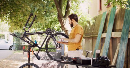 Male cyclist holding personal computer and inspects bike pedal and crank arm for damages in home yard. Active caucasian man enjoys browsing internet, fixing and maintaining modern bicycle.