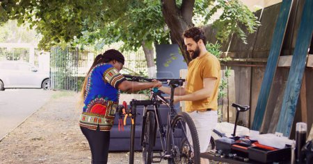 Sportbegeistertes multiethnisches Paar, das die Fahrradkarosserie zur Untersuchung und Wartung befestigt. Afrikanisch-amerikanische Freundin unterstützt Freund, indem sie modernes Fahrrad an Reparatur-Ständer im heimischen Garten klammert.