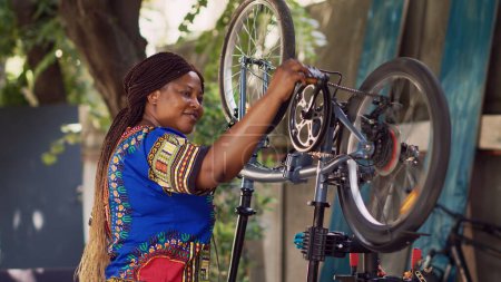 Photo for African american woman attentively checking bike components ensuring smooth and efficient summer leisure cycling. Smiling female cyclist performing bicycle maintenance. Side-view portrait shot. - Royalty Free Image
