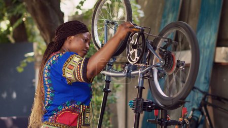 Foto de Retrato de vista lateral de la mujer negra sana deportiva que repara la bicicleta usando herramientas profesionales en el patio de casa. Mujer afroamericana ciclista evaluando y restaurando bicicleta dañada afuera. - Imagen libre de derechos
