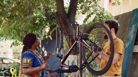 Sporty motivated couple examining and disassembling bicycle tire for maintenance with specialized equipment. Active man repairing crank arm while black woman detaches bike wheel outdoor.
