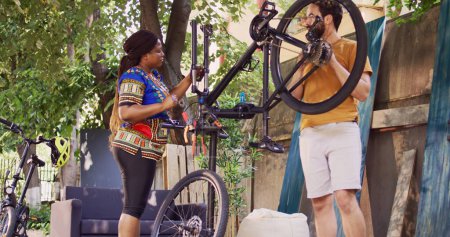 Sports-loving man examining bicycle parts in home yard while african american female cyclist removes damaged wheel. Couple performing bike maintenance outdoor with expert tools. Fade-in shot.