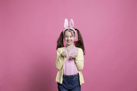 Téléchargez les photos : Joyeux jeune fille sautant autour comme un lapin devant la caméra, portant des oreilles de lapin et célébrant Pâques dimanche vacances. Petit bambin souriant riant et s'amusant sur fond rose. - en image libre de droit