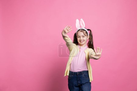 Photo for Little schoolgirl smiling and posing in front of the camera, cheerful small child thinking about gifts on easter holiday celebration. Young cute child wearing bunny ears over pink background. - Royalty Free Image
