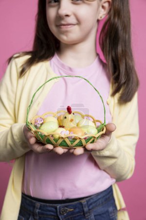 Photo for Beautiful small girl with bunny ears holding an egg basket for easter festivity, posing with pretty spring trinkets. Happy and energetic child celebrating the festive season in the studio. - Royalty Free Image
