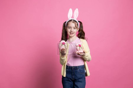 Foto de Niña con orejas de conejo sosteniendo decoraciones de Pascua rosa delante de la cámara, presentando su huevo hecho a mano y conejo relleno. Niño pequeño sonriendo en el estudio y celebrando la fiesta de primavera. - Imagen libre de derechos