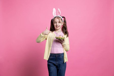 Téléchargez les photos : Sur fond rose, une jolie petite fille attrape un arrangement d'œufs et une poulette comme articles de Pâques. Petit dépeignant l'excitation à l'idée des célébrations du printemps et de la jeunesse, oreilles de lapin. - en image libre de droit