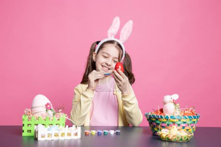 Photo for Little enthusiastic child drawing colorful designs and painting eggs with watercolors to prepare for the Easter festivities, pink backdrop. Young small girl relaxing as she creates art at table. - Royalty Free Image