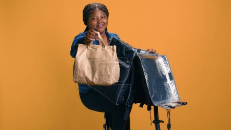 Mujer joven y saludable negro da pulgares hacia arriba mientras entrega comida para llevar asegurando la satisfacción del cliente. Repartidor afroamericano que ofrece alternativa ecológica para cenar en el vecindario.