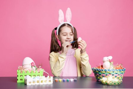 Photo for Small cheerful toddler decorating eggs with watercolor for easter holiday preparations, coloring festive ornaments against pink background. Young little girl having fun with art and craft. - Royalty Free Image