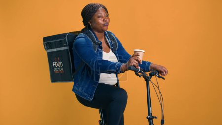 Afroamerikanerin auf dem Fahrrad genießt eine Tasse Kaffee, bevor sie ihren rasanten Lieferservice-Job verrichtet. Junge Kurierin entspannt sich nach Paketzustellung auf Abruf.