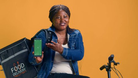 Afrikanische Kurierin, die ein digitales Gerät mit grünem Bildschirm hält, während sie auf dem Fahrrad sitzt. Junge Zustellerin zeigt Smartphone mit leerer Kopiervorlage.